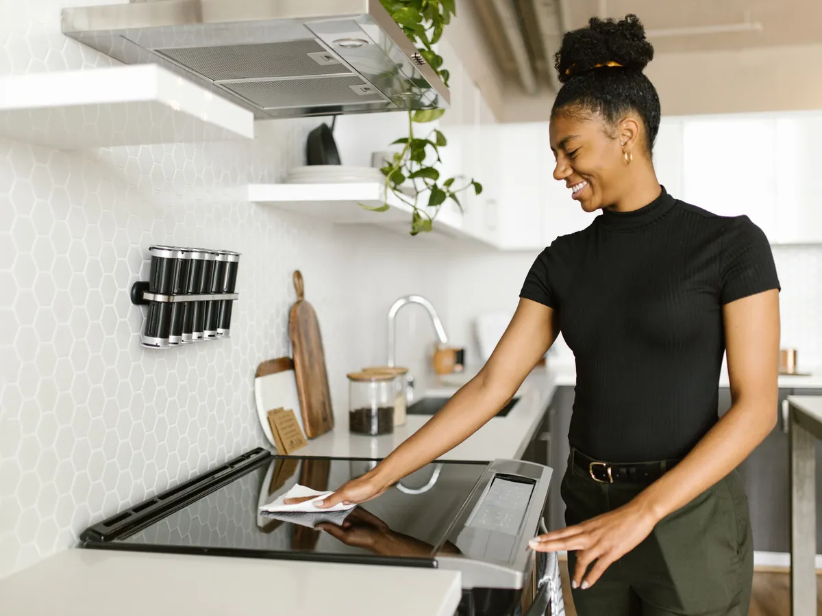 Smiling cleaner wiping a glass stovetop in a spotless white kitchen.